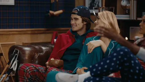 A woman in teal pajamas leans over to kiss a man in a red hoodie on the cheek while friends relax beside them.