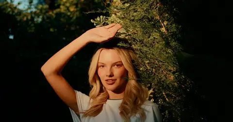A blonde woman shades her face with one hand in warm light beside dense desert greenery.