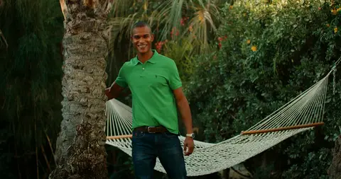 A man in a green polo stands beside a white hammock in a leafy resort garden.