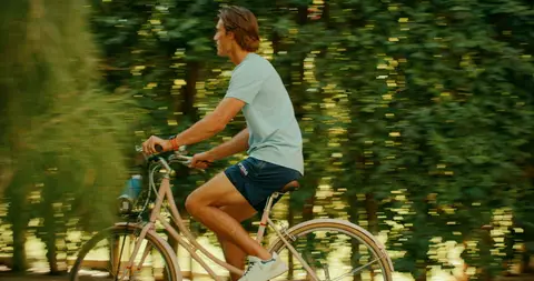 A cyclist in a white polo and denim shorts rides a bicycle through sunlit greenery.