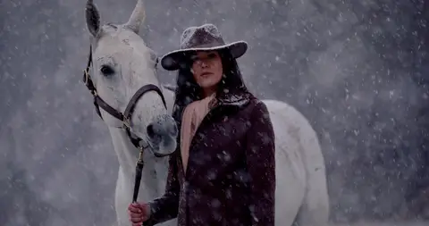 A woman in a wide-brim hat leads a white horse through heavy falling snow.