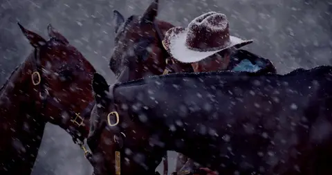Several dark horses cluster around a bundled handler during heavy snowfall.