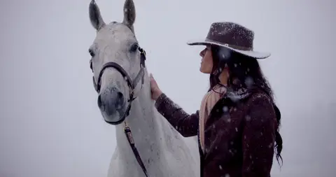 A woman in a wide-brim hat touches a white horse against a pale snowy background.