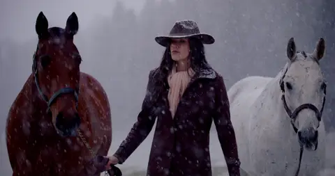 A woman stands between a dark horse and a white horse as snow falls around them.