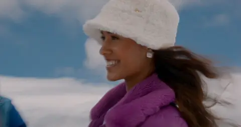 A smiling woman in a purple coat and white fuzzy hat turns against a snowy mountain sky.