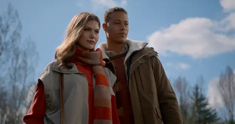 A woman and man pose together in layered coats and scarves beneath a blue sky.