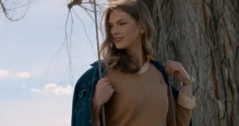 A woman in a blue coat stands beside a tree beneath a pale winter sky.