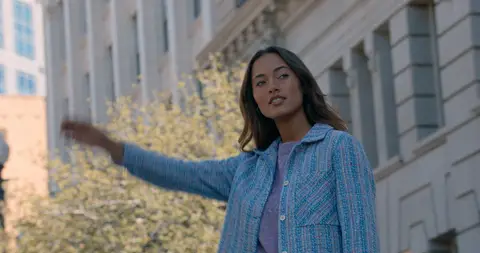 A woman gestures while wearing a blue tweed jacket beside a light stone building.