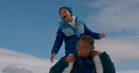 A laughing woman rides on a man's shoulders beneath a bright sky in a snowy mountain setting.