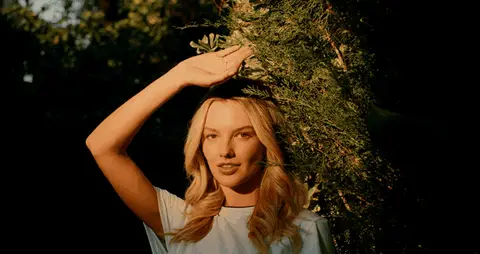 A blonde woman raises her hand against the sun while posing beside desert greenery.