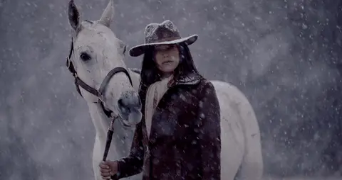 A woman leads a white horse as dark horses pass through dense falling snow.