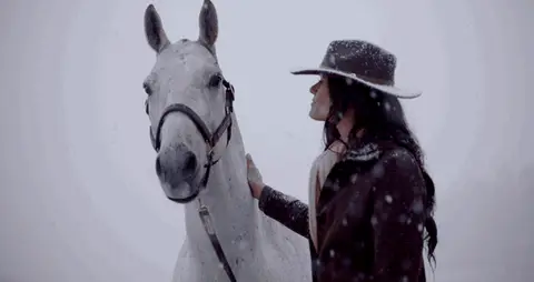A woman moves among several horses before the group walks across a snowy field.