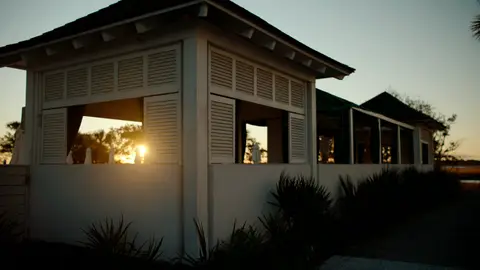 The setting sun shines through an open coastal pavilion beside silhouetted buildings.