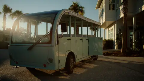 A pale vintage-style shuttle is parked beside a coastal house in warm backlight.