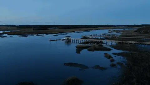 A slow aerial approach moves across blue-hour water toward a long dock and marsh islands.